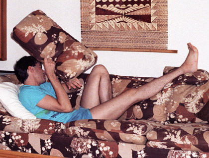 Dave laying on a couch with his foot propped up while hiding his face from the camera and wearing impossible short turquoise shorts.