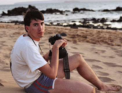 Dave sitting on a Hawaiian beach with his camera looking disappointed and wearing shorts that are too short.