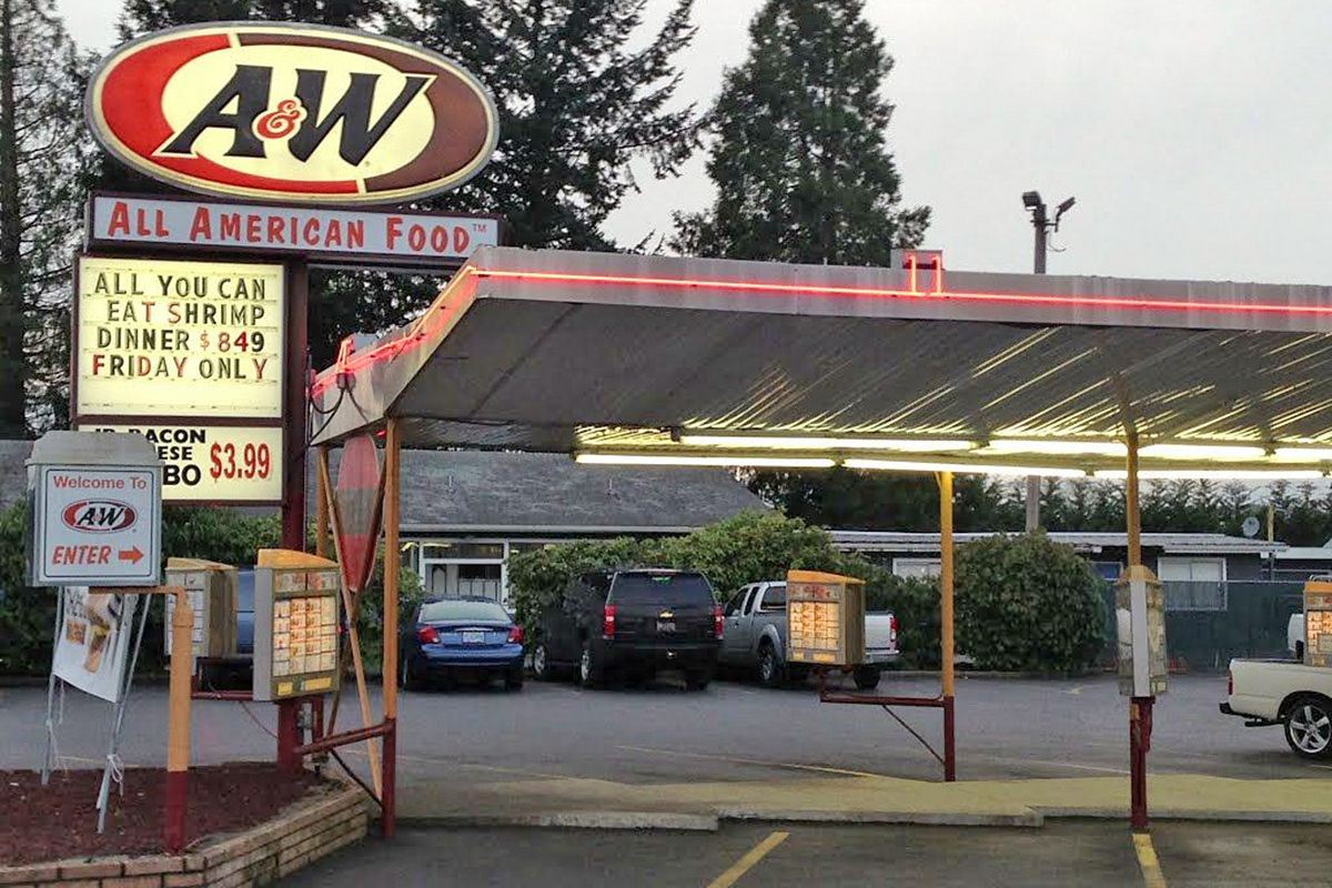 A photo of an old A&W drive-in at dusk showing parking spots with the car-hop menu boards sticking out and a sign saying ALL YOU CAN EAT SHRIMP DINNER $849 FRIDAY ONLY on an illuminated billboard attached to the road-sign.