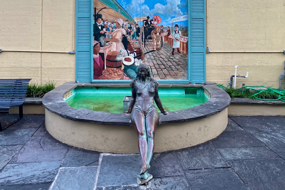 Statue of a woman in brass sitting on the edge of a fountain with a painting of New Orleans behind it.