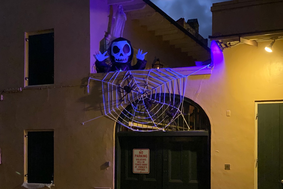 A yellow building with a spooky purple glow coming from the balcony where a skeleton monster is peeking out and there’s a spider in a giant web hanging over.