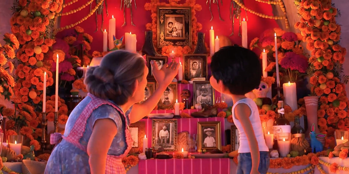 A frame from the animated masterpiece COCO from Disney Pixar showing a colorful altar decorated with marigold flowers, candles, and photos of the dead while a grandmother is explaining it to her grandson Miguel.