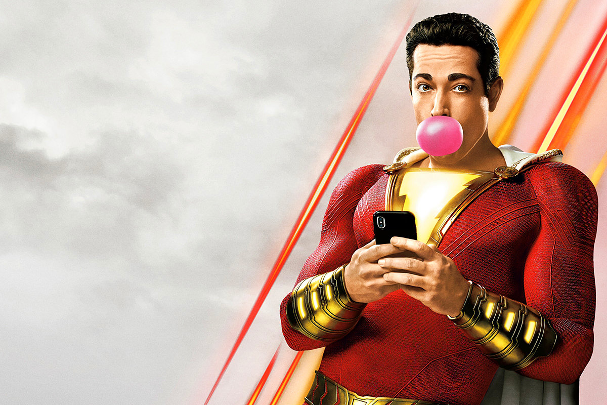 Shazam in his stupid suit looking at a stupid mobile phone while stupidly blowing a bubble with some gum.