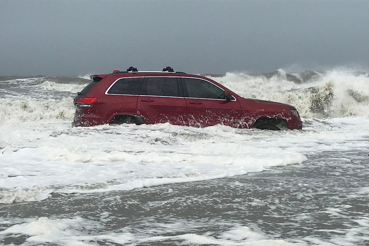 A photo of a red SUV stuck on the beach while waves crash around it.