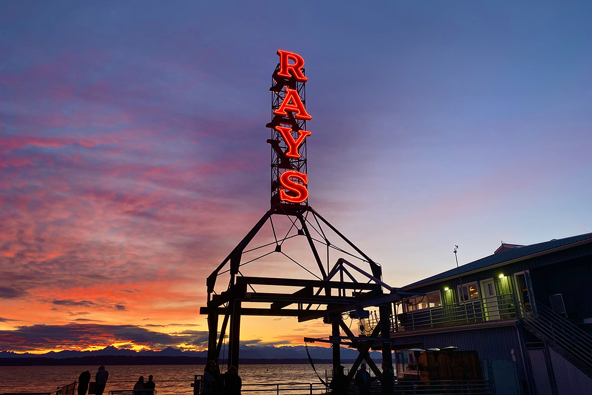 A beautiful shot of the Ray's Boathouse neon sign glowing while intense colors of sundown light up the sky behind it.