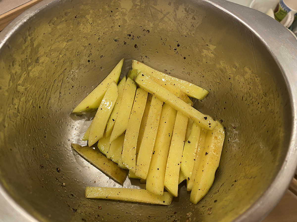 Potatoes sliced into fry planks in a metal bowl... they are oiled and covered with seasoning salt and pepper.