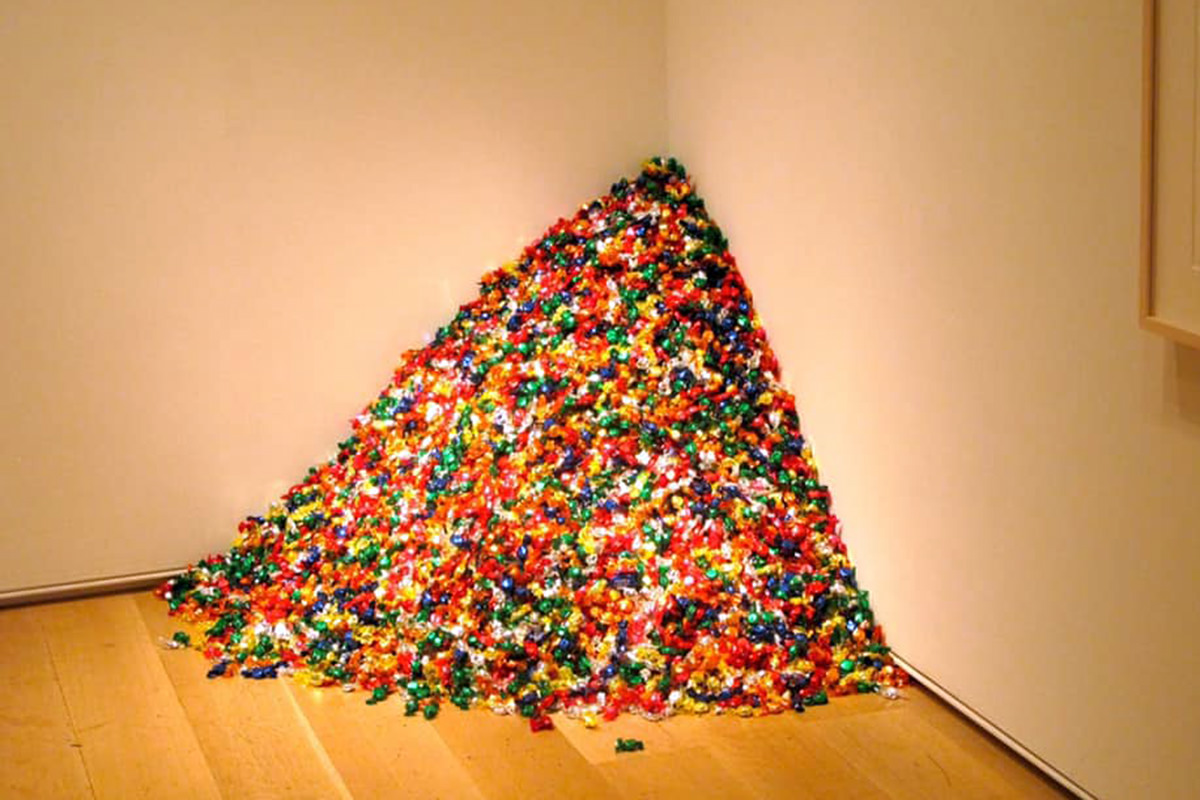 A corner in a white-walled art gallery. There is a large mound of multicolored candy piled up in the corner.