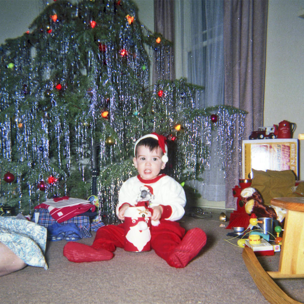 Young me sitting in front of a Christmas tree with Santa pajamas, complete with hat.