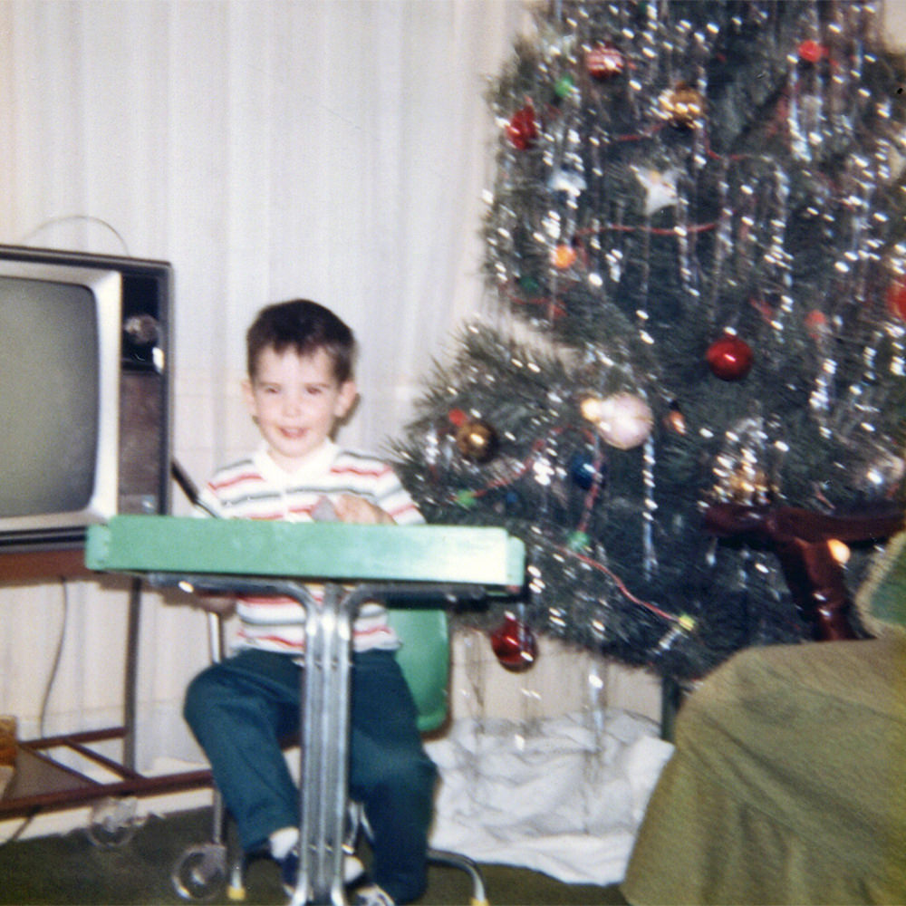 Young me sitting in a desk in front of a Christmas tree.