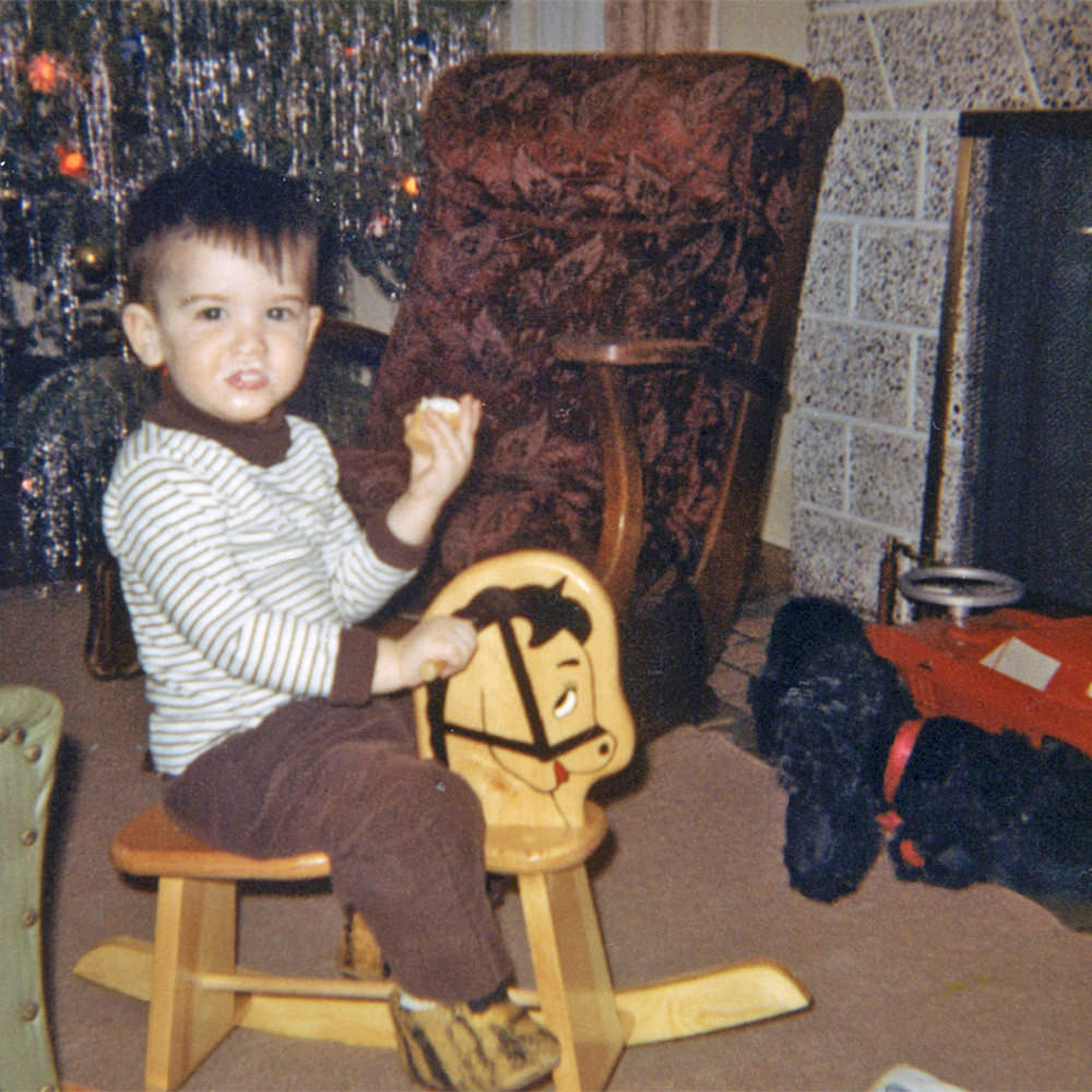 Young me sitting on a small rocking horse in front of a Christmas tree.