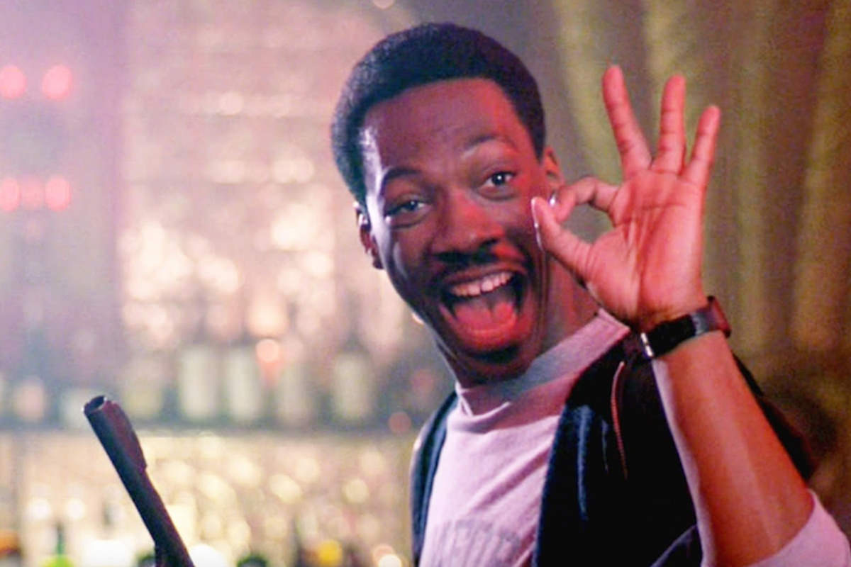 Eddie Murphy as Axel Foley from Beverly Hills Cop making the OKAY sign in one hand while holding a gun in the other.