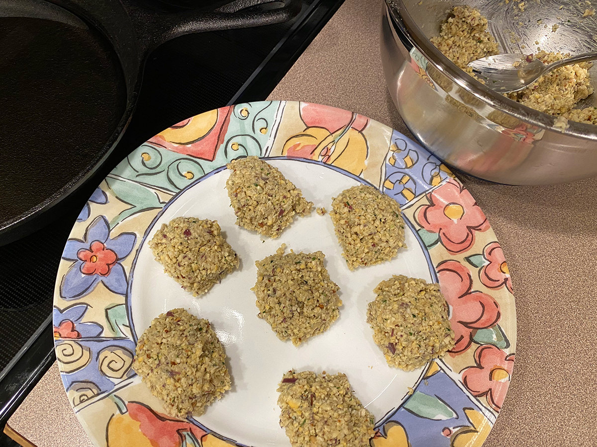 Freshly-made falafel patties sitting on a plate next to a metal bowl filled with falafel mix and a skillet of hot oil.