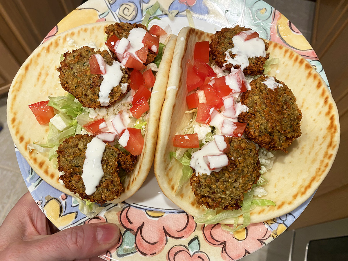 Finished falafel patties on top of a flatbread with shredded lettuce, tomato, onion, and red pepper with a cream sauce drizzled on top.
