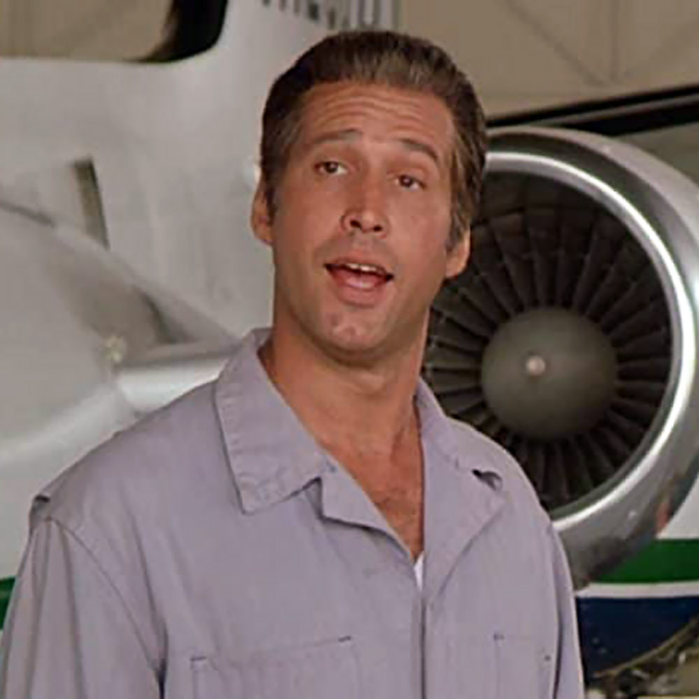 Chevy Chase as G. Gordon Liddy from the movie Fletch Lives standing in front of an airplane and dressed as a flight mechanic.