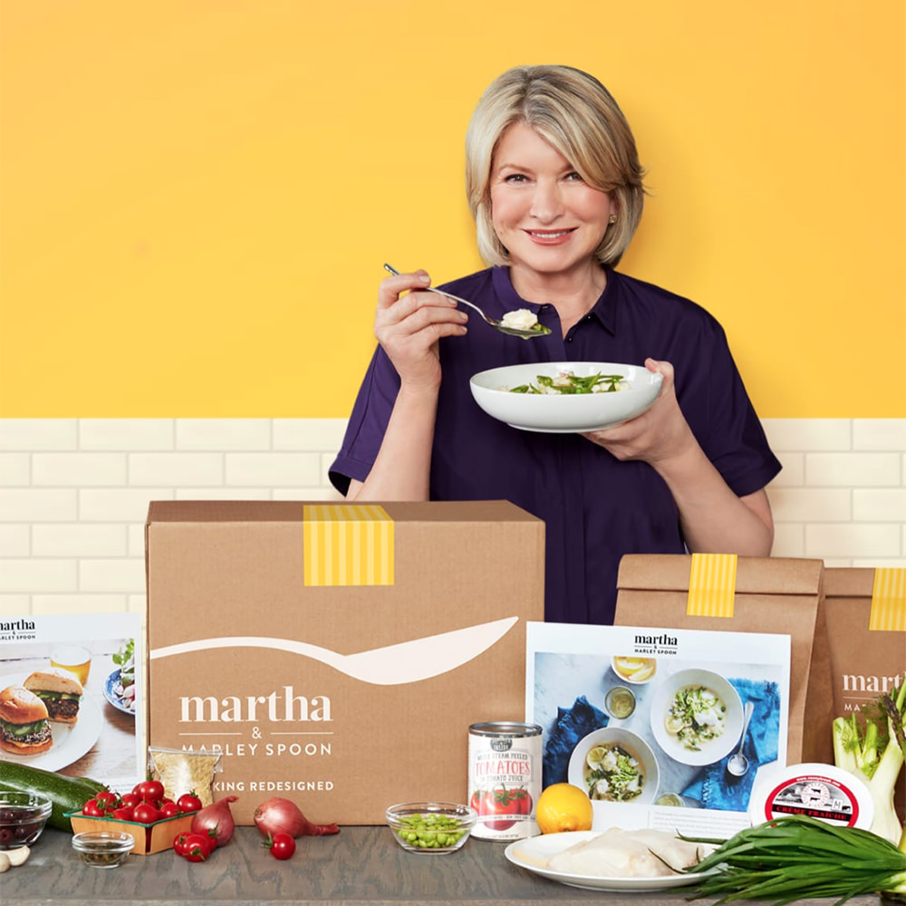 Martha Stewart eating a bowl of food in front of boxes of Marley Spoon stuff with ingredients in front.