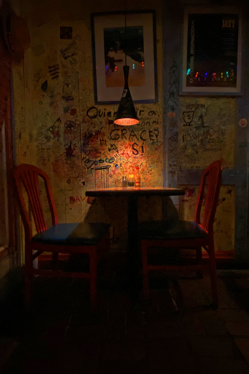 In a darkened corner of a small restaurant, a light above a small table illuminates grafitti-strewn walls as two brilliant red chairs sit empty.