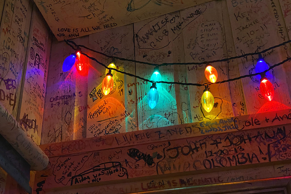 A ceiling corner of the restaurant with grafitti covering every possible surface. Colorful Christmas bulbs glow in the darkness, illuminating phrases such as LOVE IS ALL WE NEED.