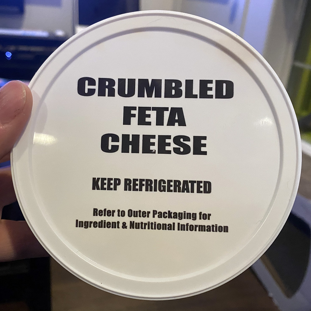 The lid of a tub of feta cheese which says CRUMBLED FETA CHEESE - KEEP REFRIGERATED on top in generic black lettering.