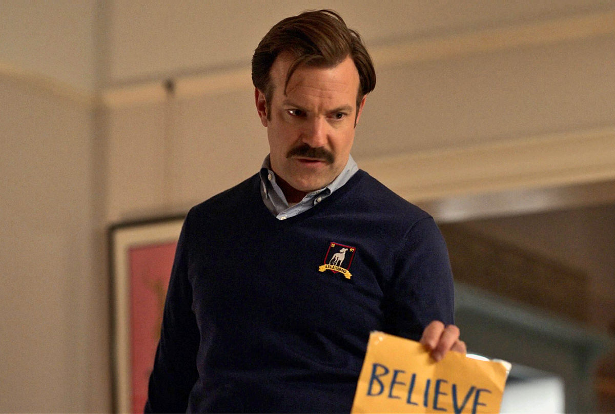 Ted Lasso holding up a handwritten sign that says BELIEVE.