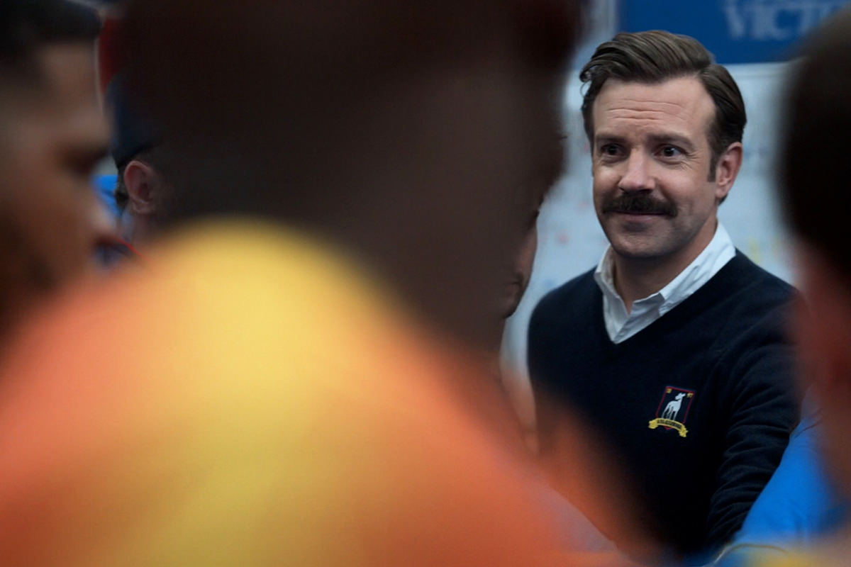 Ted Lasso smiling in a crowded locker room.