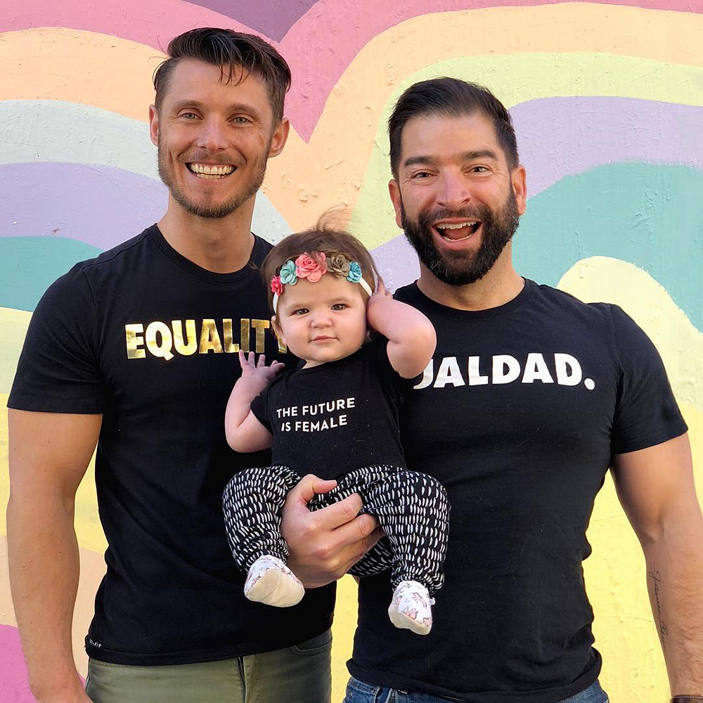 Two dads with their baby girl... who is wearing a T-shirt which says THE FUTURE IS FEMALE.