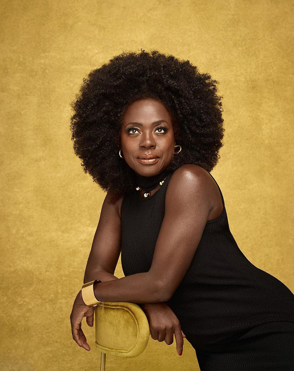 Viola Davis dressed in black shot in front of a gold background looking gorgeous as she looks upward.