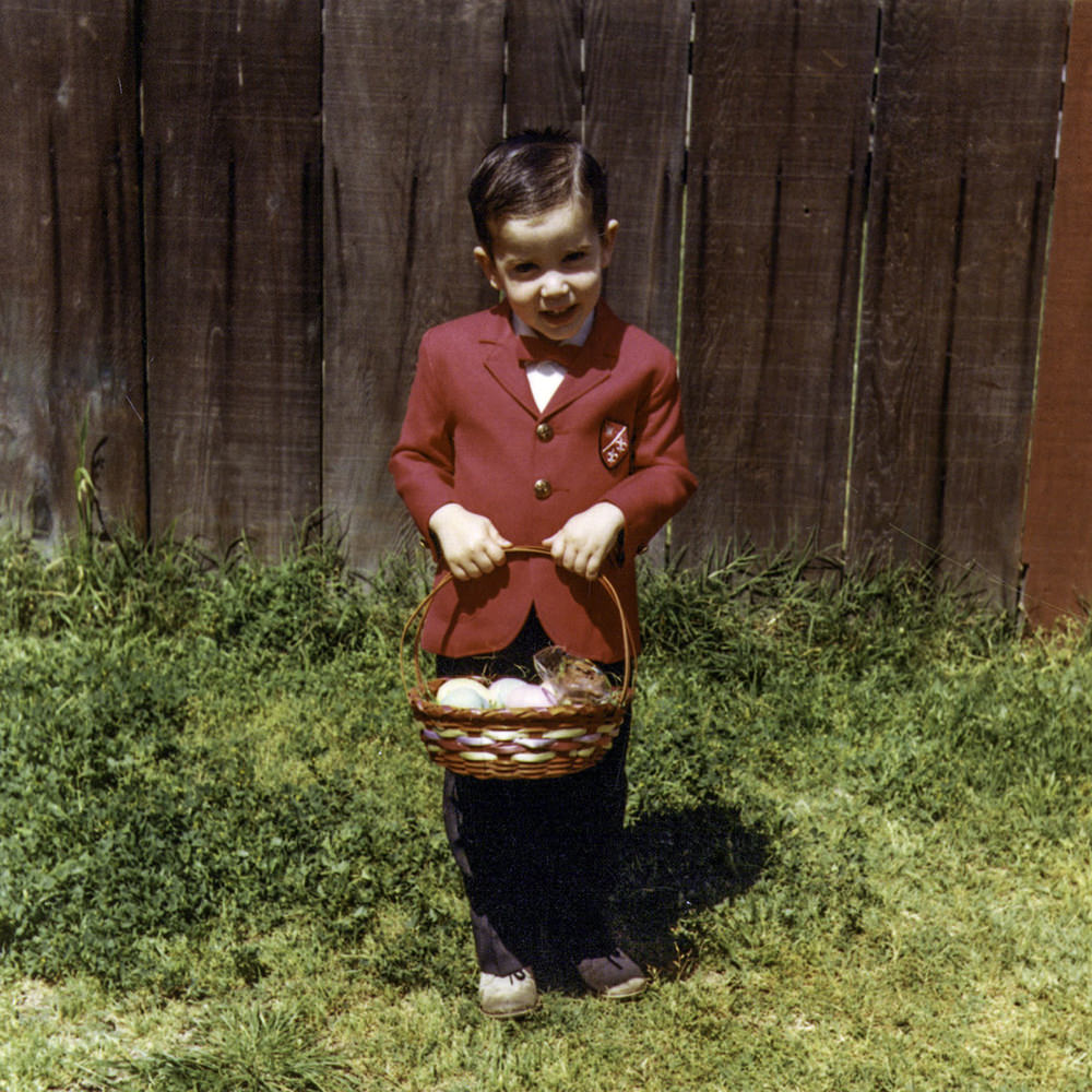 Young me with an Easter basket in the back yard with green grass and a fence behind me.