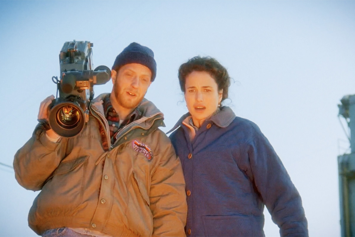 Andie MacDowell and Chris Elliot looking down the cliff at the explosion, which is reflecting on them with an orange glow.