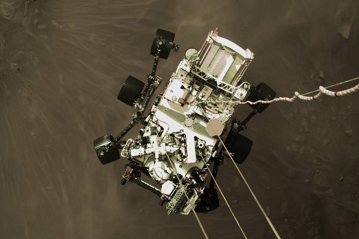 Photo of the Perseverance rover being lowered to Mars from the viewpoint of the lander module looking down at it.