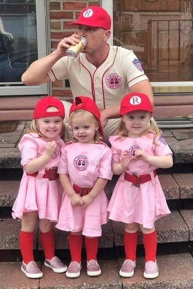 Three young girls dressed as players from A League of Their Own while a man behind them drinks a beer in his coaches uniferm.