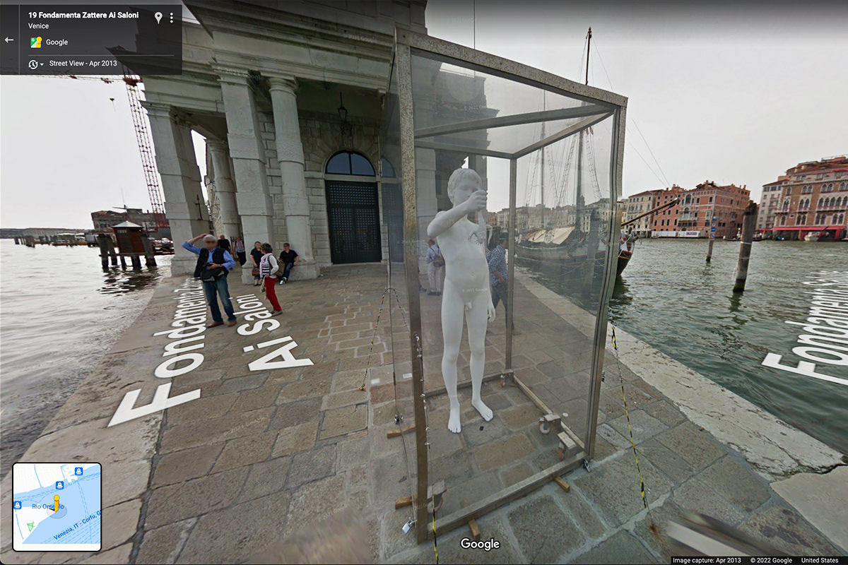 A Google Maps Street View capture of a large white statue of a boy holding a frog which has been encased in a glass cage.