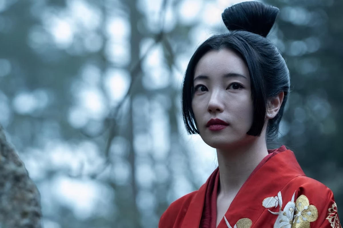 A Japanese woman stands in a bleak forest with her red kimono jumping out of the shot.