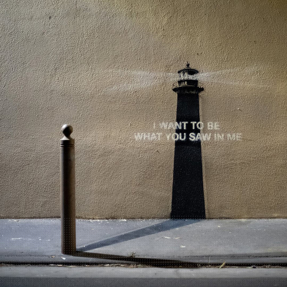 A stencil lighthouse painted on a wall in black shadow with I WANT TO BE WHAT YOU SAW IN ME spraypainted in white over it.